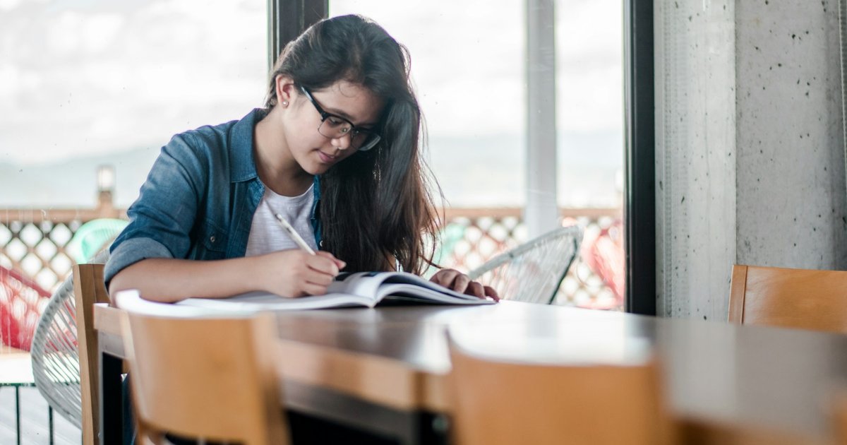 Student in glasses writing notes into an open book at a table by a large window