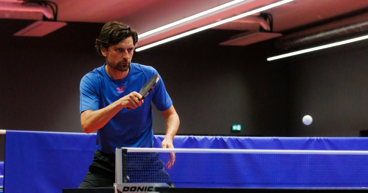 A man in a blue shirt returning a table tennis ball across a blue table under bright lights
