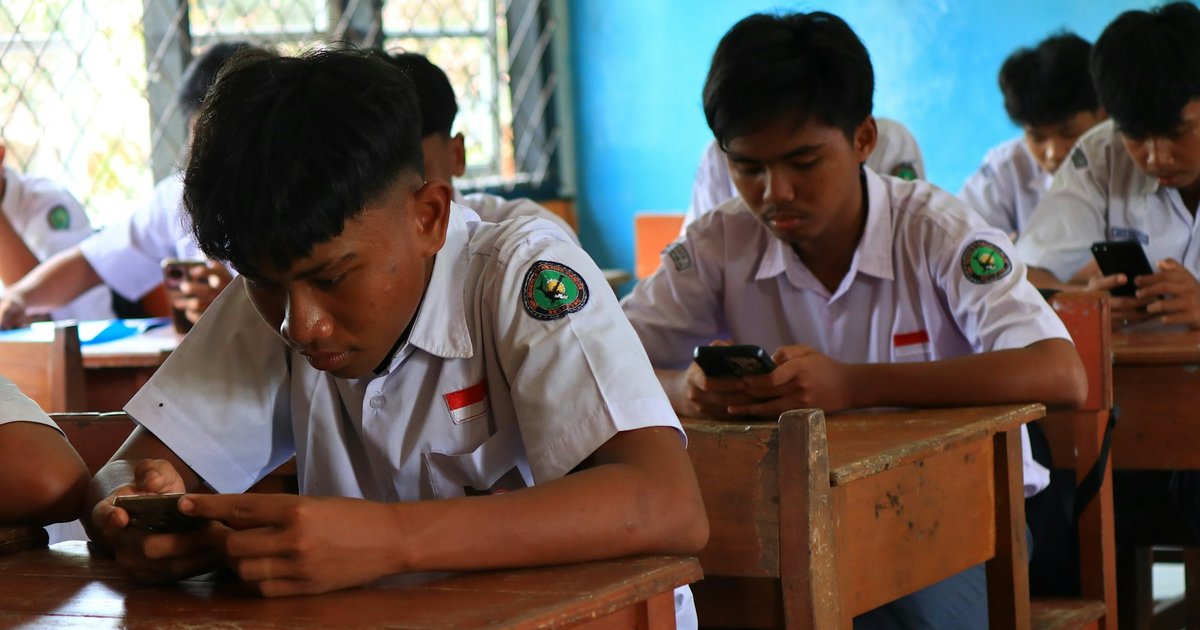 Secondary school students in white uniforms answering on their phones at classroom desks