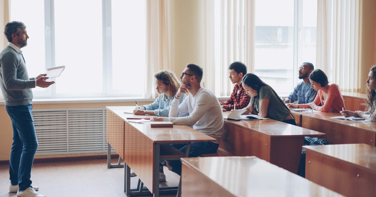 Teacher holding a tablet in front of a bright classroom while students sit at wooden desks