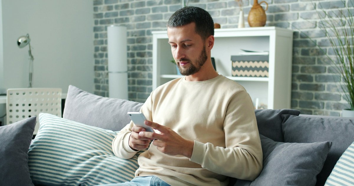 Man sitting on a grey sofa at home looking down at the smartphone in his hands