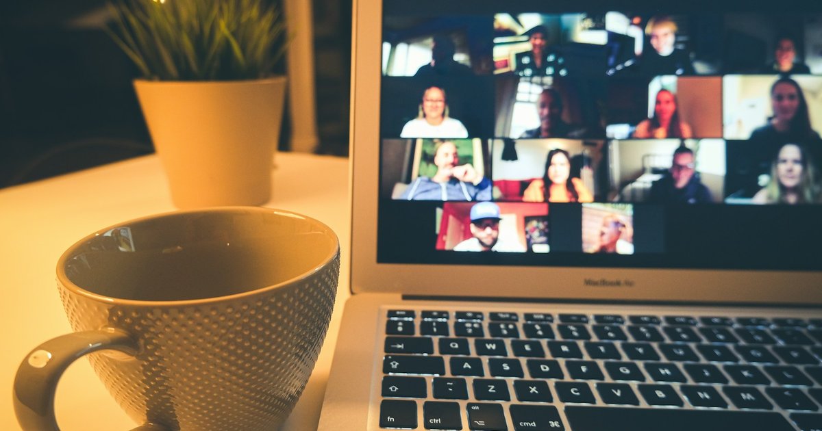 Laptop on a table showing a group video call with many participants, next to a coffee mug and a small plant